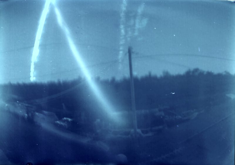 Cyanotype-toned solargraph aiming east showing bright arcing Sun trails over a silhouetted landscape with cylindrical pinhole distortion