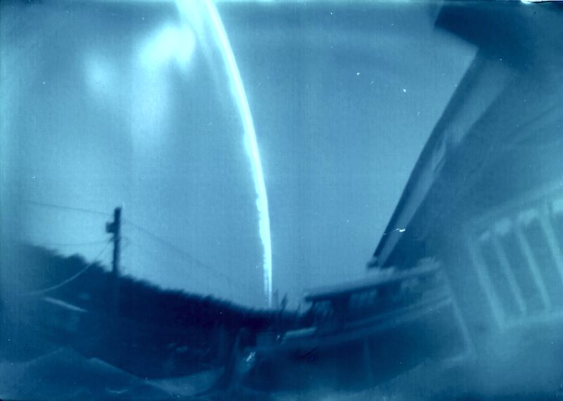 Cyanotype-toned solargraph aiming west with a vertical Sun arc, utility pole silhouette, and curved pinhole horizon